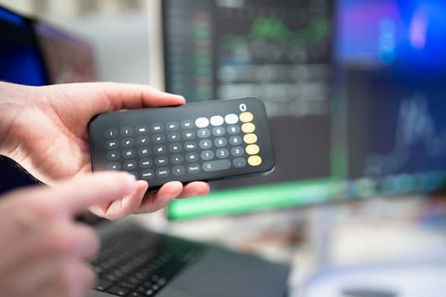 Eine Hand hält einen Taschenrechner. Im Hintergrund sieht man verpixelte Bildschirme.
